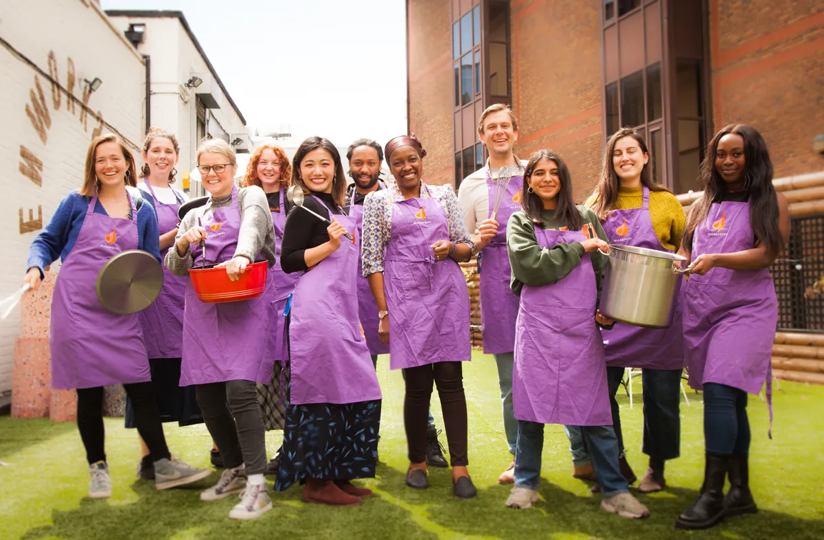 A group of people in purple aprons participating in a Migrateful London Cookery Class, posing for a picture.