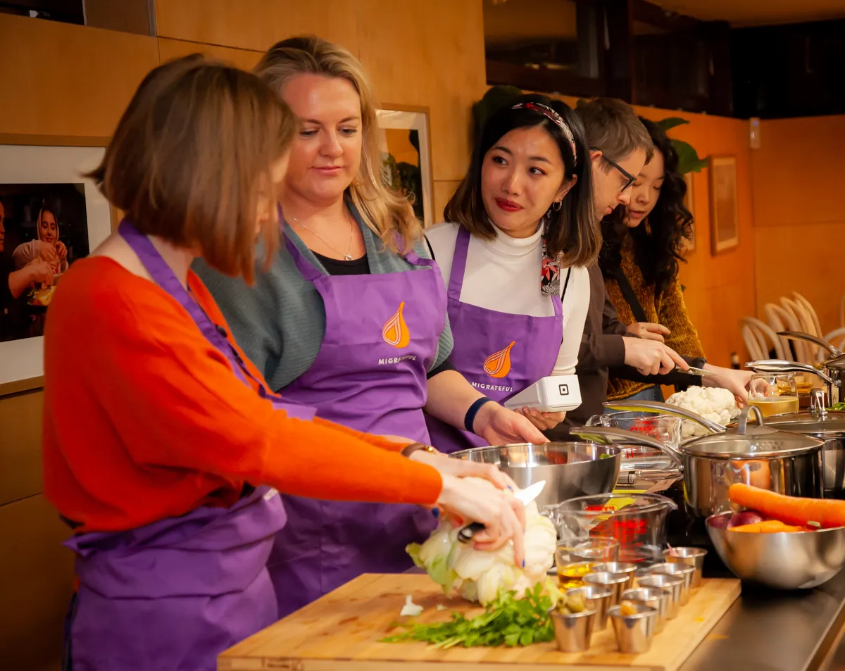 A group of women in purple aprons cooking together at a Migrateful London Cookery Class.