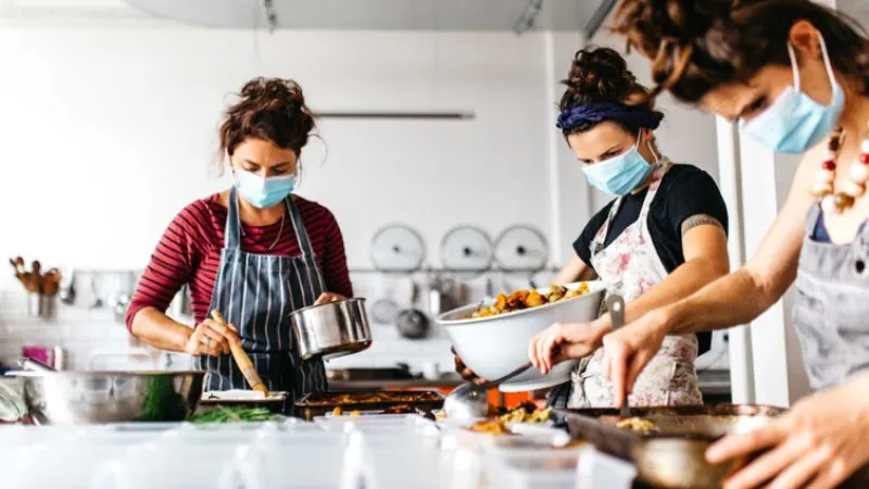 Migrateful women in face masks preparing food in a kitchen.