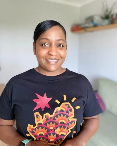 A woman wearing a black t-shirt standing in a living room, excitedly preparing for her upcoming London Cookery Class organized by Migrateful.