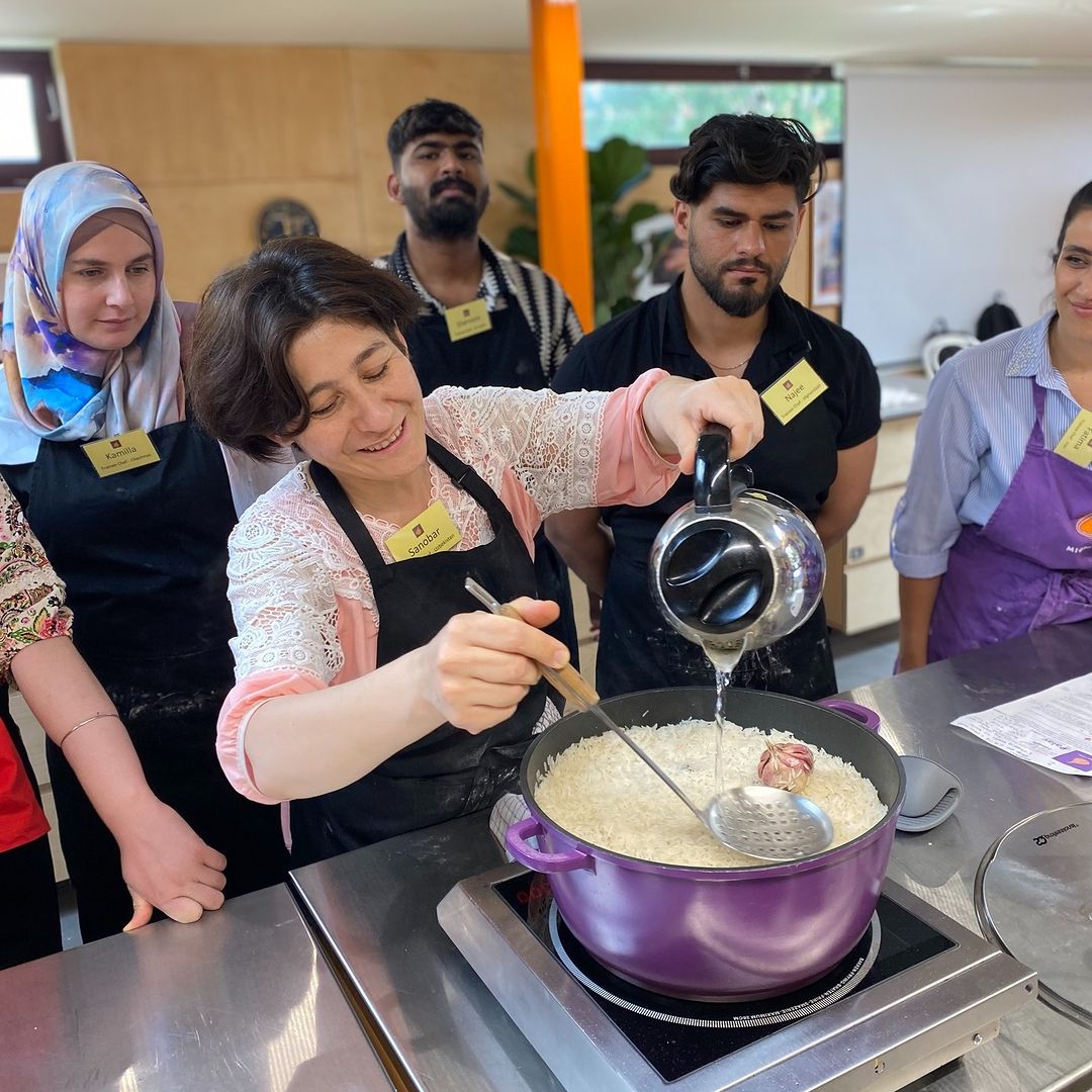 A woman stands cooking over a pot while others watch