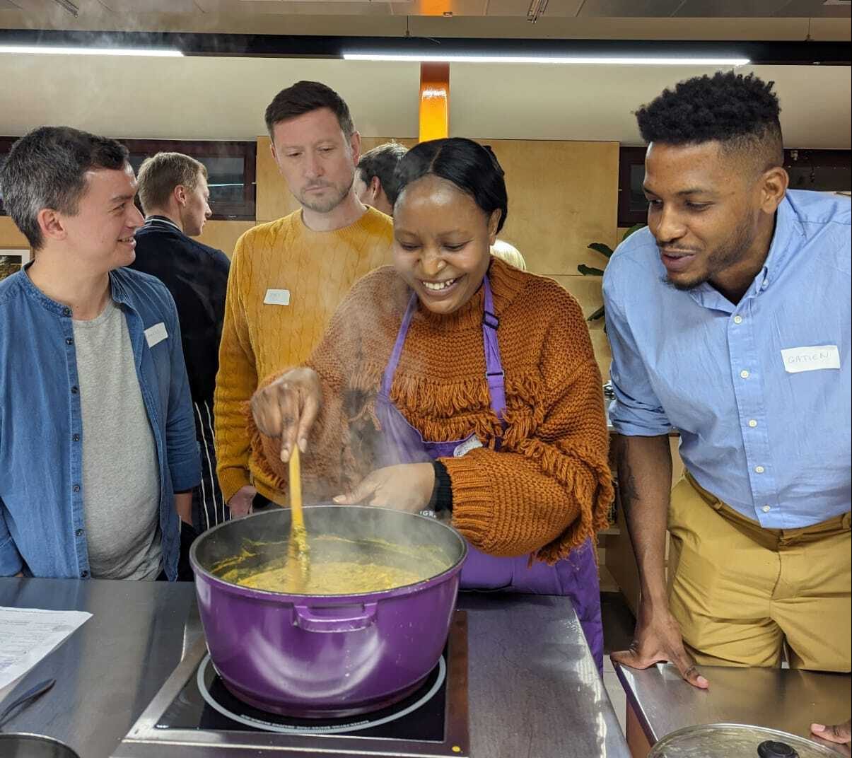 Three people look into a pot of food, while a fourth stirs it with a spoon.