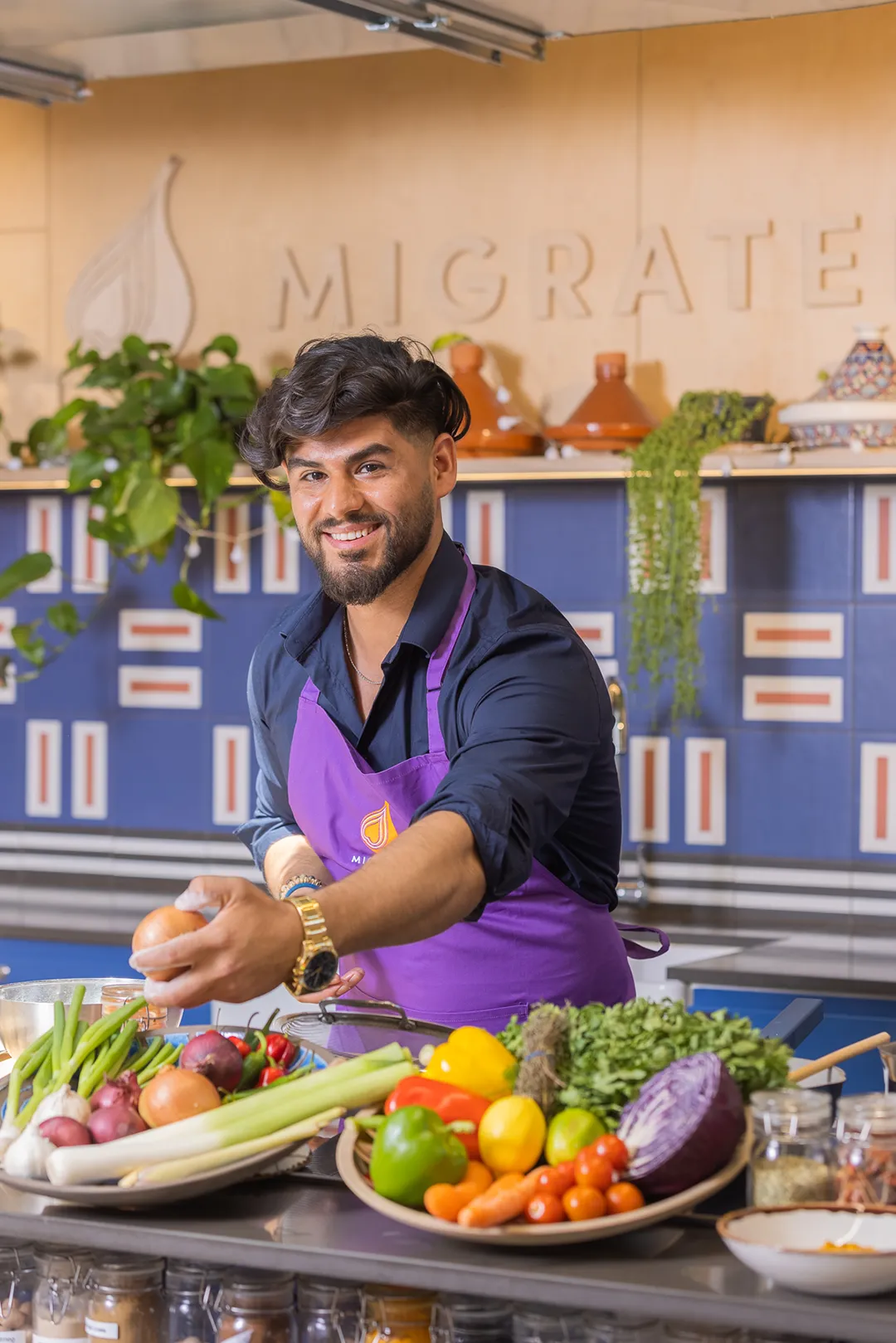 A man in an apron standing in front of a kitchen full of vegetables, participating in a London Cookery Class.