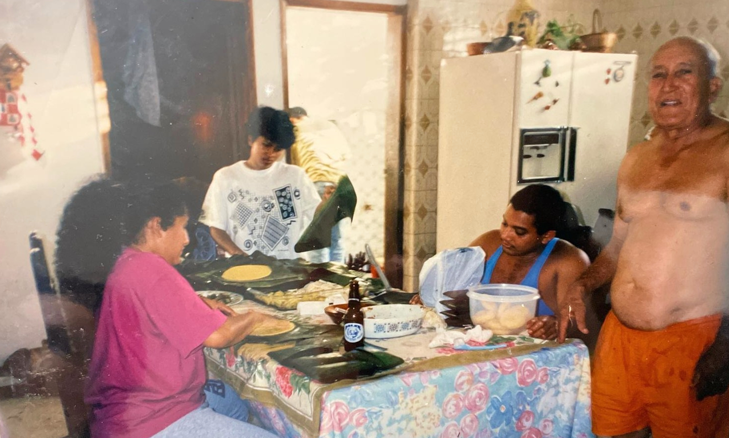 A family gathered around a table with a floral tablecloth, preparing Hallacas. An elderly man stands on the right.