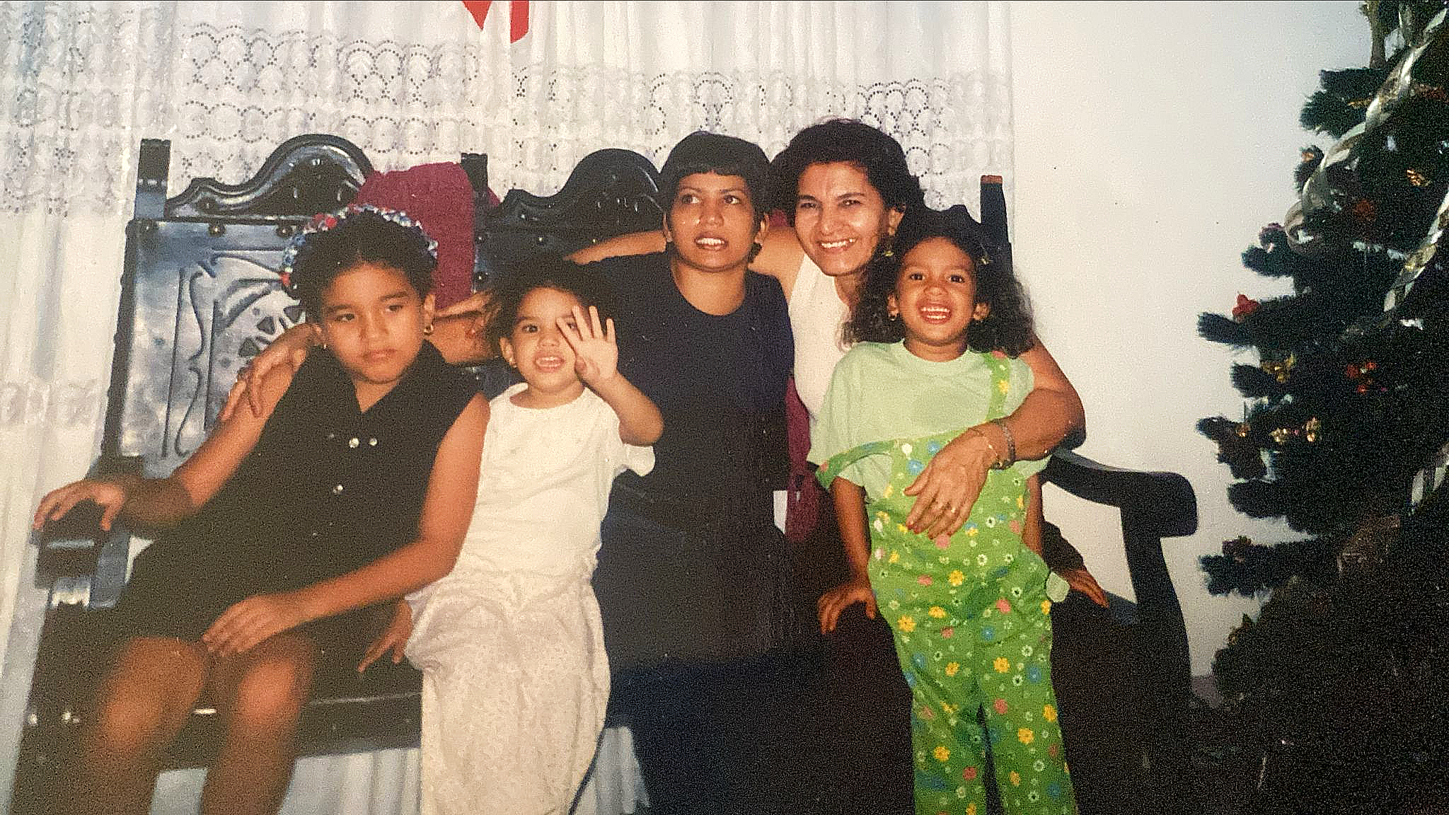 A group of two adult women and three kids smiling sitting on a sofa, and posing together for a photo. A partially visible Christmas tree stands on the right.